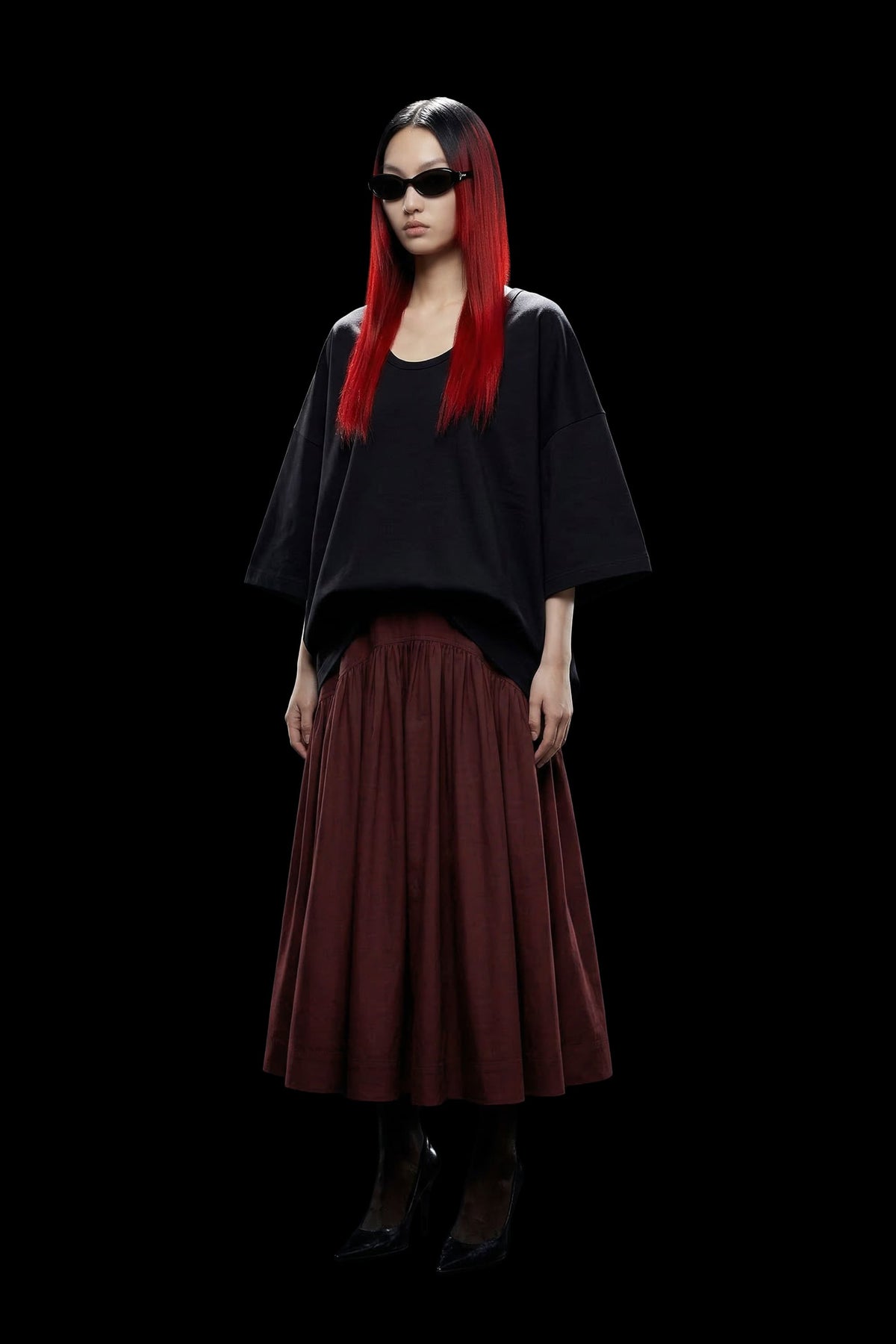 A model with long, straight red hair and black sunglasses is shown from the waist down, wearing a loose black top and a pleated dark red skirt, set against a solid black background.