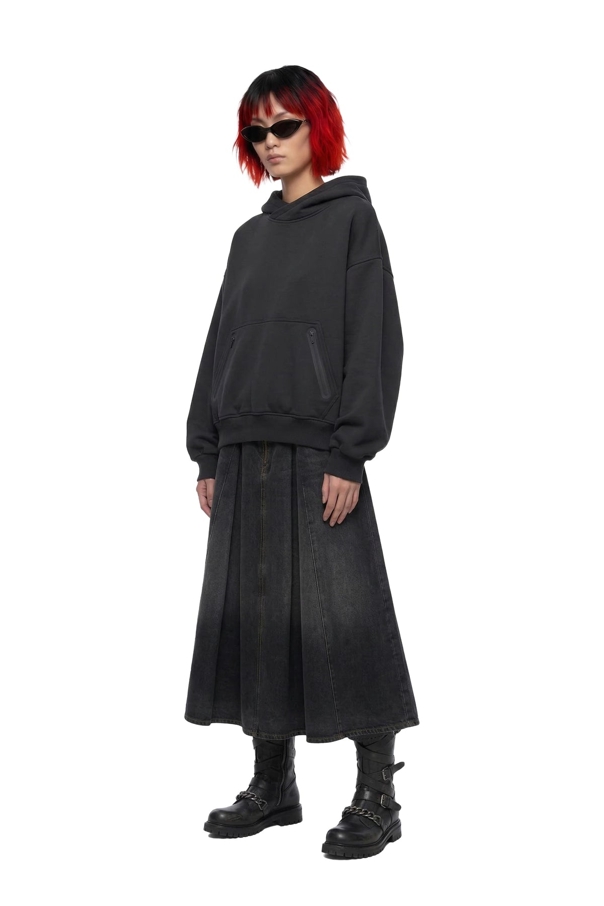 A model with short red hair and black sunglasses wears a black oversized hooded sweatshirt, a long black pleated skirt, and black combat boots against a plain white background.