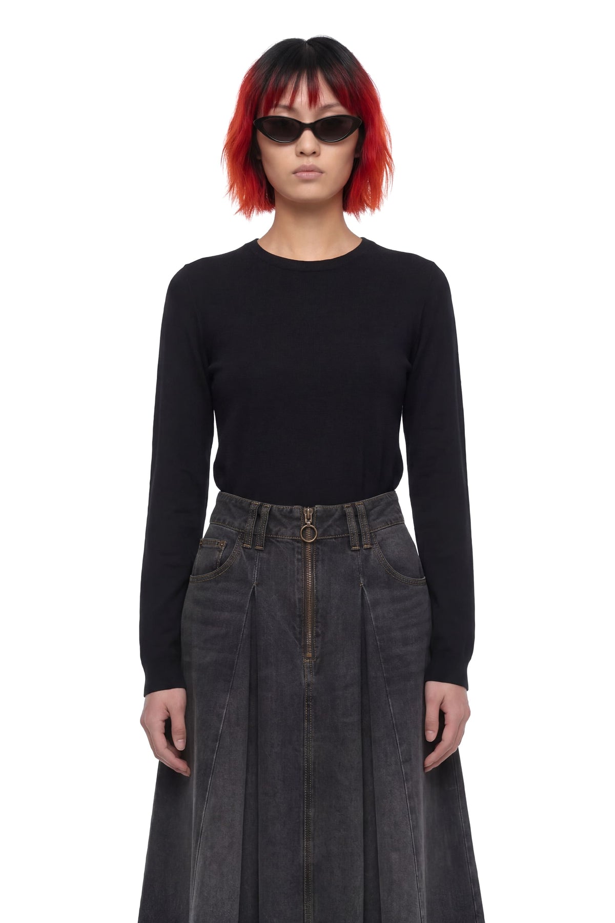 A model with short red and black hair wearing a black long-sleeve crewneck top and a dark gray pleated denim skirt, standing against a plain white background.