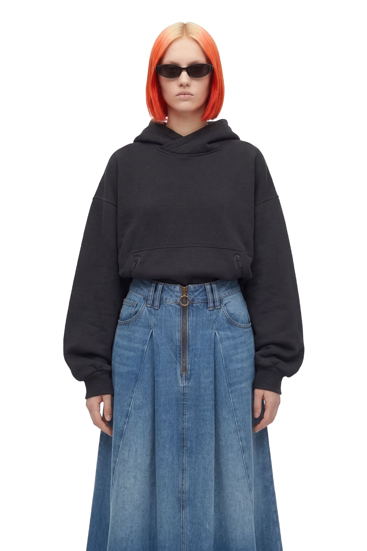 A model with bright orange hair and black sunglasses wears a black cropped hoodie and a high-waisted blue denim skirt with a front zipper, shot against a solid white background.