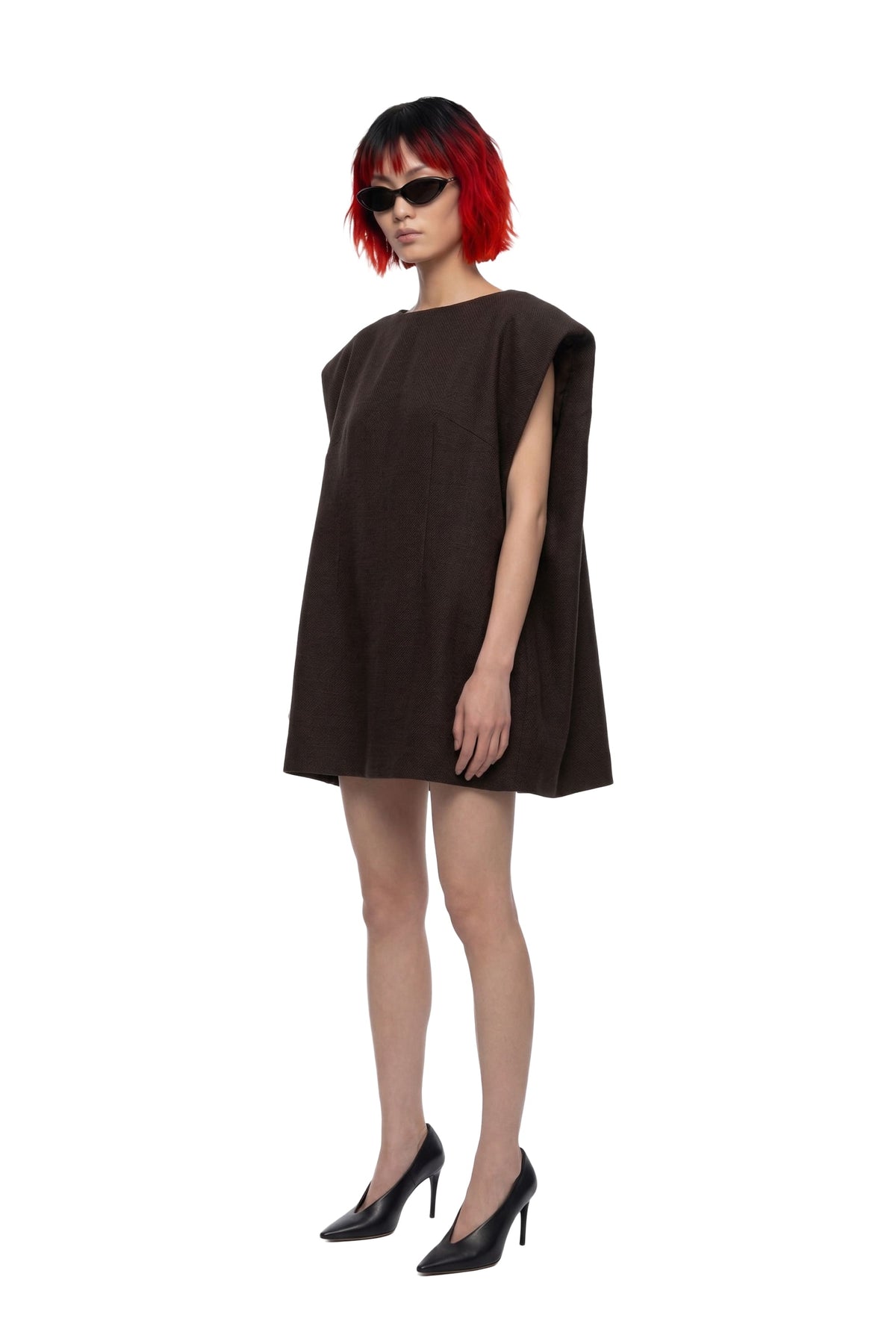 A model with short red hair and black sunglasses wearing a dark brown sleeveless mini dress and black heels, standing against a plain white background.