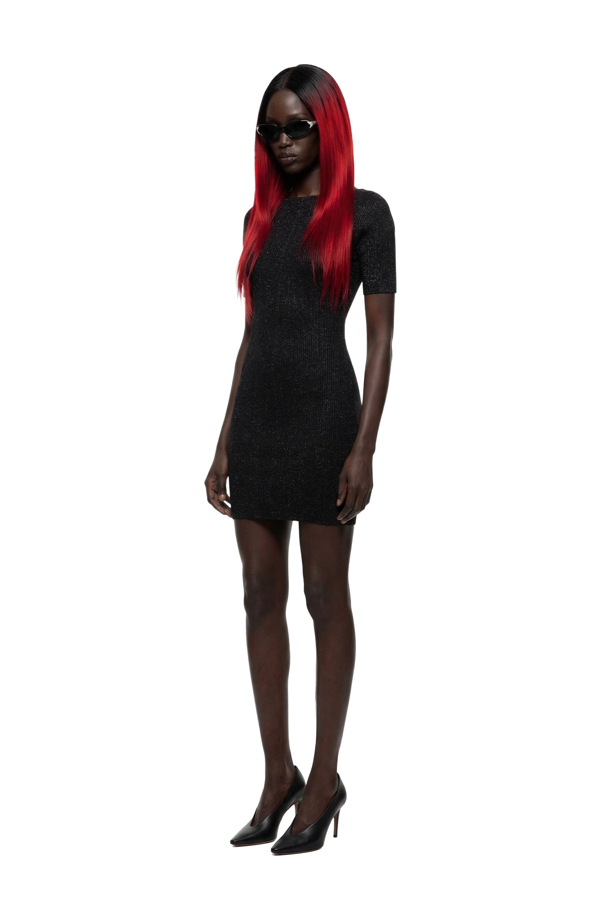 A model with long red hair and black sunglasses wearing a short-sleeved black textured mini dress and black heels, standing against a solid white background.