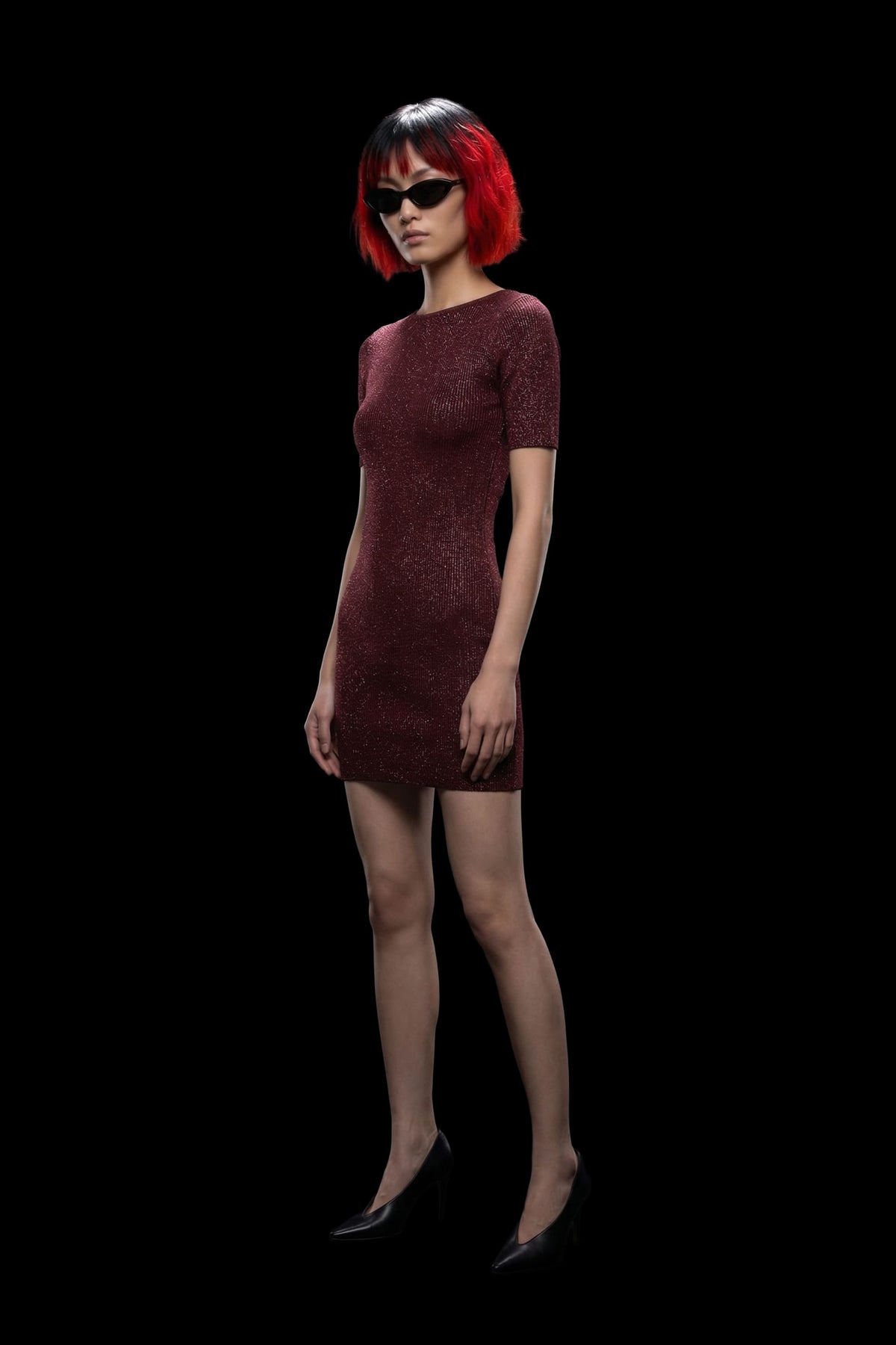 A model with a short red and black bob haircut and black sunglasses wears a short-sleeved, textured burgundy mini dress and black heels, standing against a solid black background.