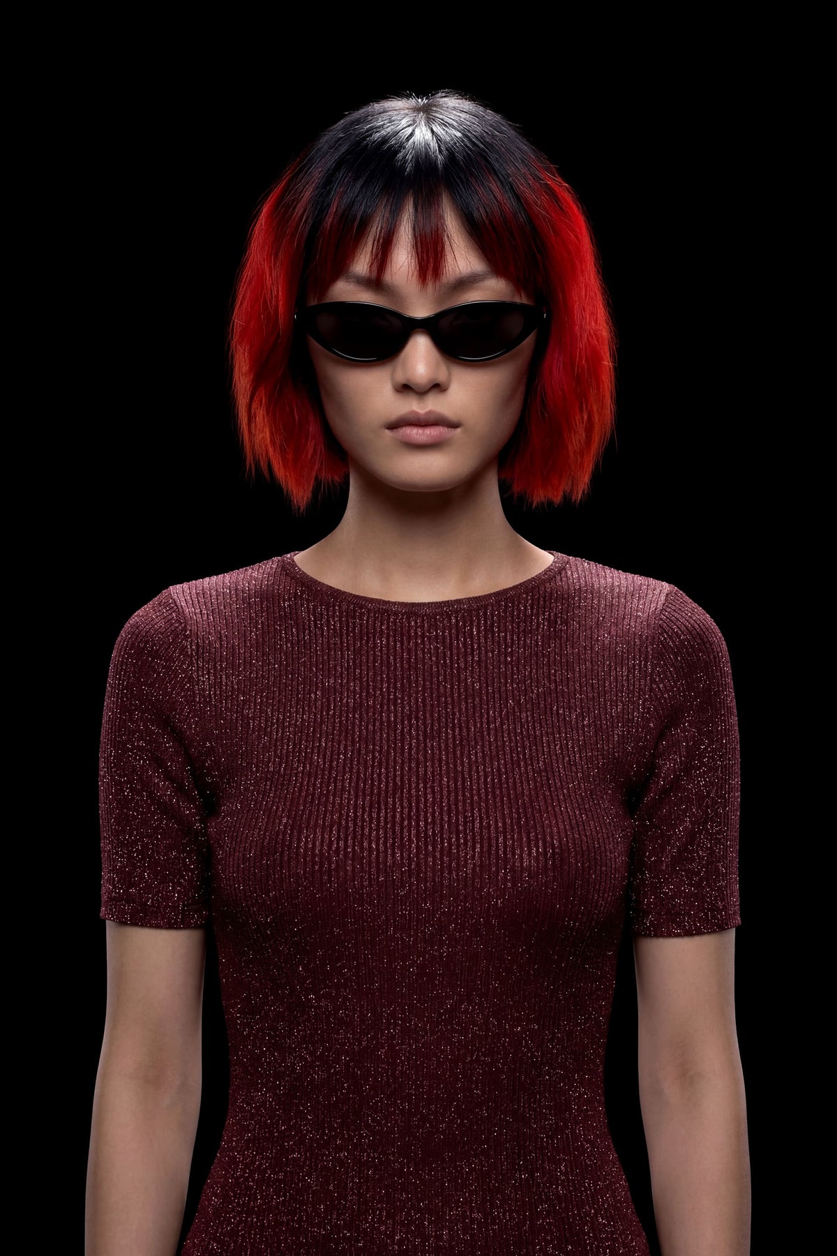 A model with a short black and vibrant red ombre bob hairstyle wearing black cat-eye sunglasses and a sparkling dark burgundy short-sleeve top, set against a solid black background.
