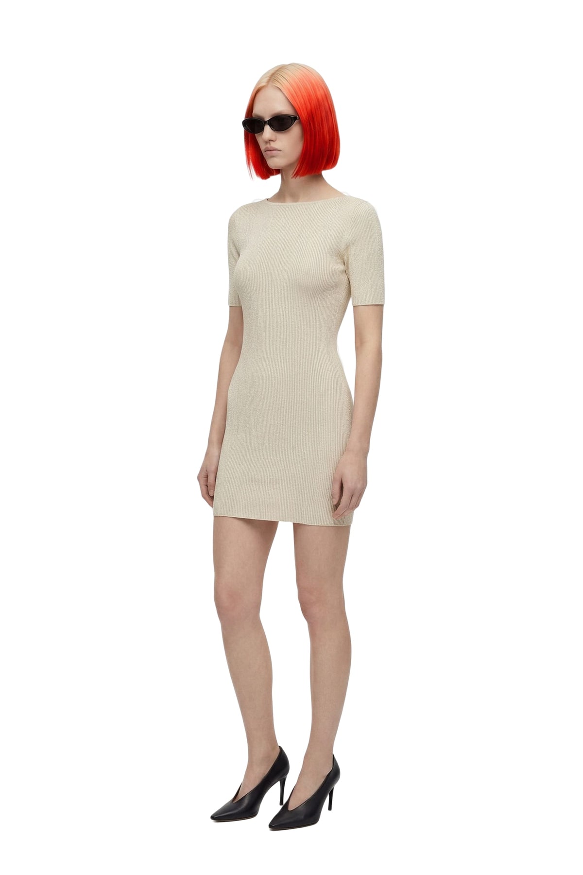 A model with a vibrant orange-red bob haircut and black sunglasses wears a short-sleeve beige knit mini dress with black high heels, set against a plain white background.
