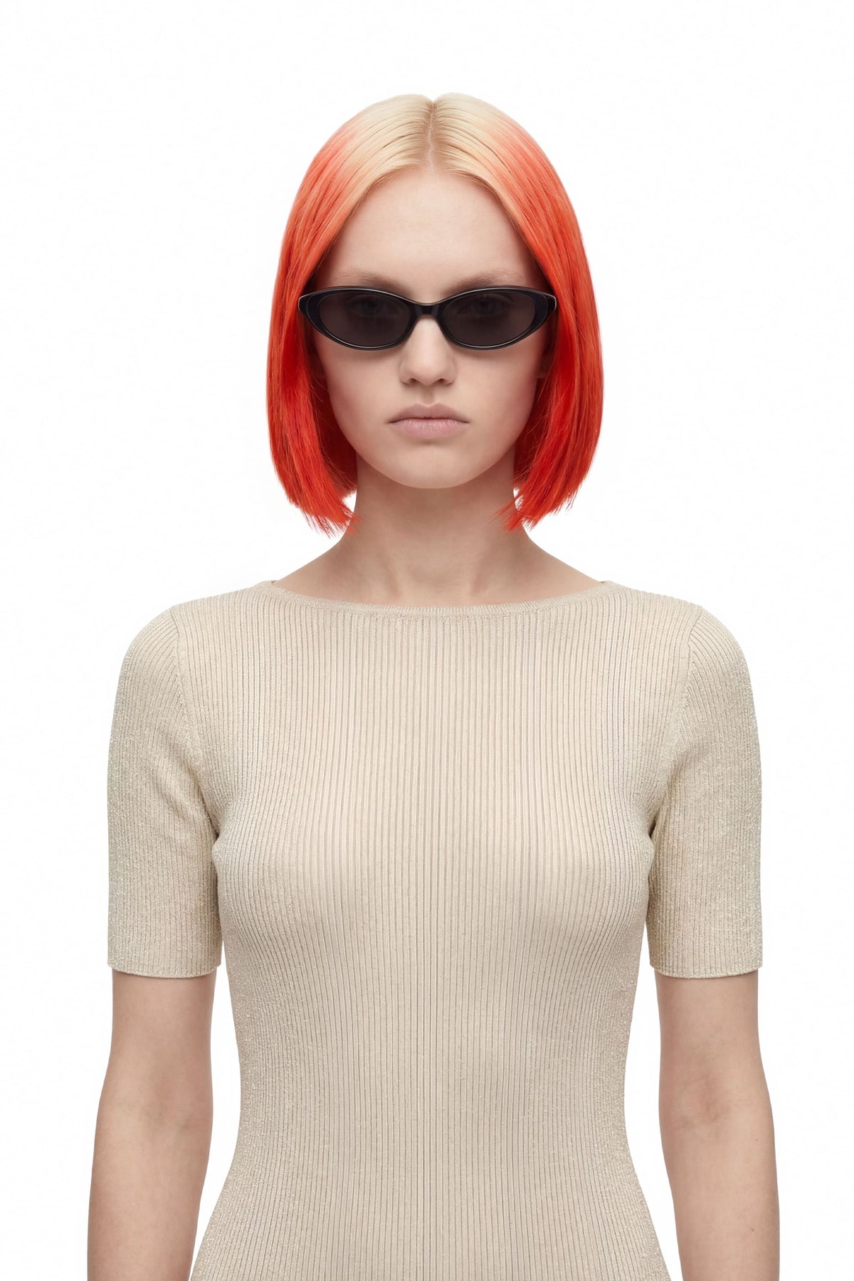 A model with a short, bright orange-red ombre bob haircut wearing black cat-eye sunglasses and a beige ribbed short-sleeve top, photographed against a solid white background.