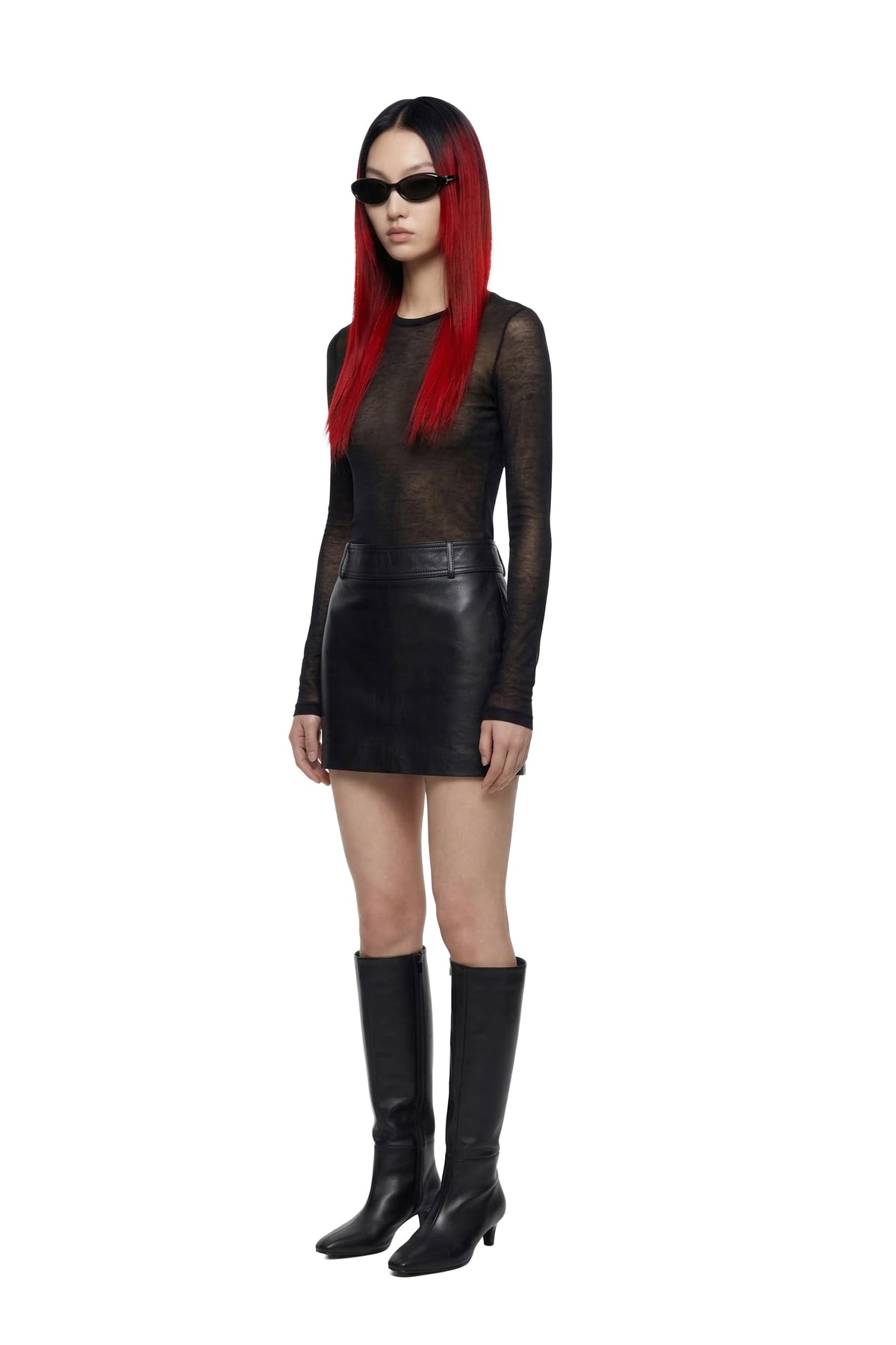 A model with long red hair and black sunglasses is wearing a black sheer long-sleeve top, a black faux-leather mini skirt, and black knee-high boots, standing against a solid white background.