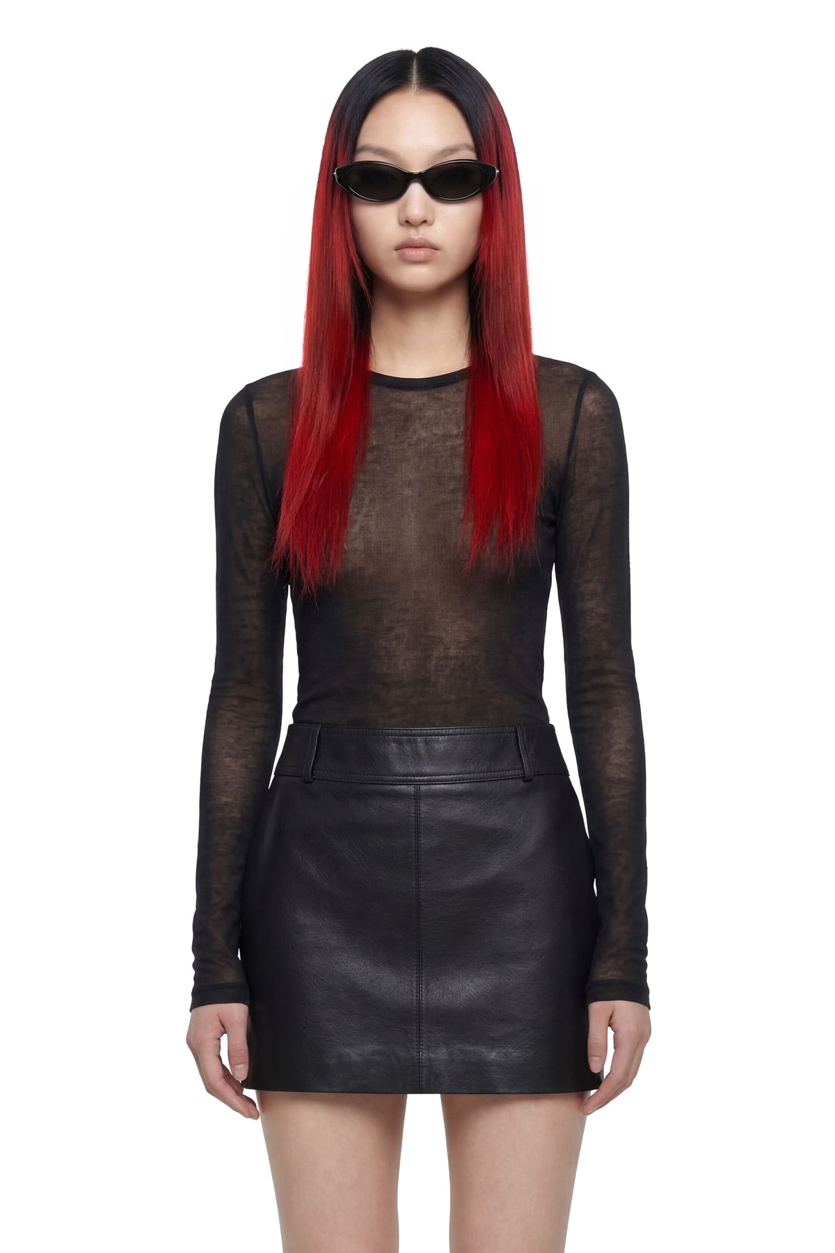 A model with long red hair wearing a sheer black long-sleeve mesh top and a black faux-leather mini skirt, styled with black sunglasses, against a plain white background.