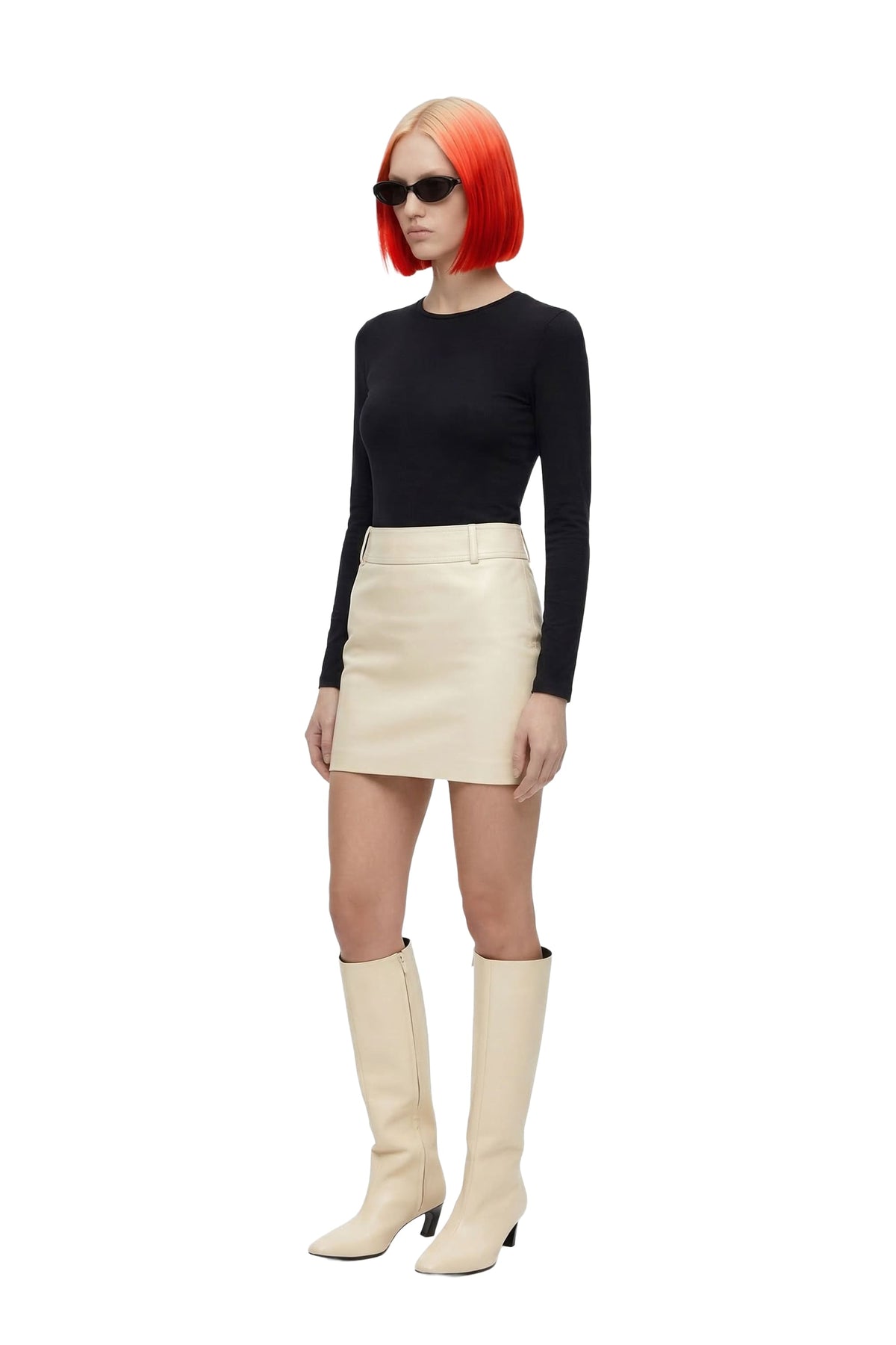 A model with a bright orange-red bob haircut wearing a black long-sleeve top, a cream-colored mini skirt, and matching cream knee-high boots, set against a plain white background.