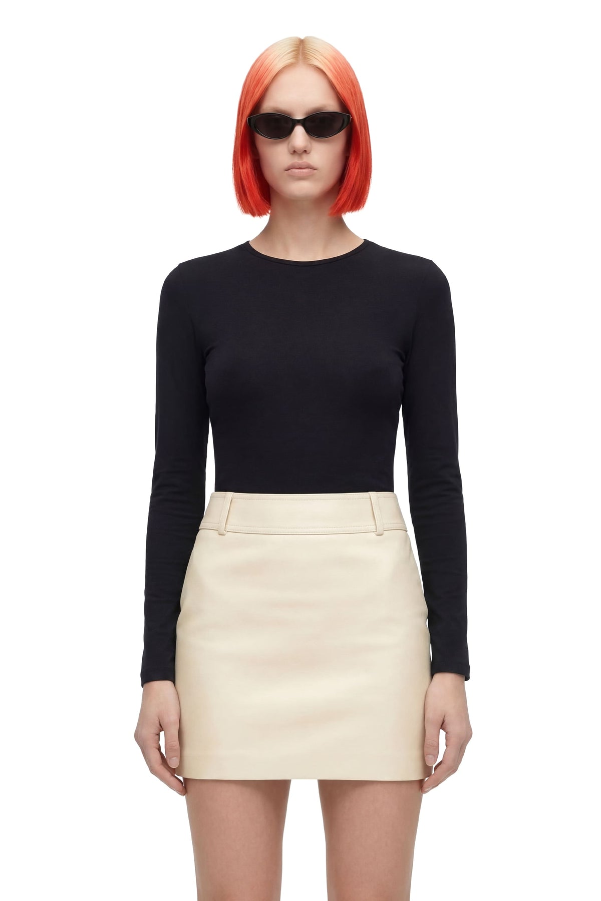 A model with bright orange hair and black sunglasses, wearing a black long-sleeve crewneck top and a cream-colored mini skirt, photographed against a solid white background.