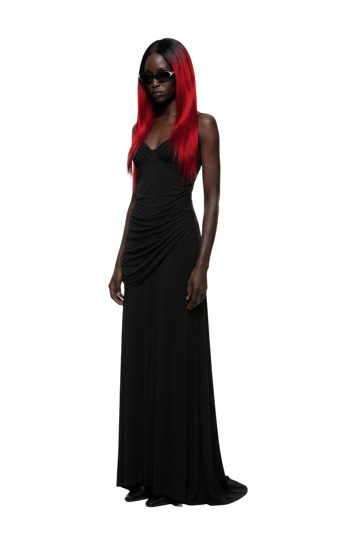 A woman modeling a floor-length black gown with draped detailing on the bodice and skirt, paired with black sunglasses, against a plain white background.
