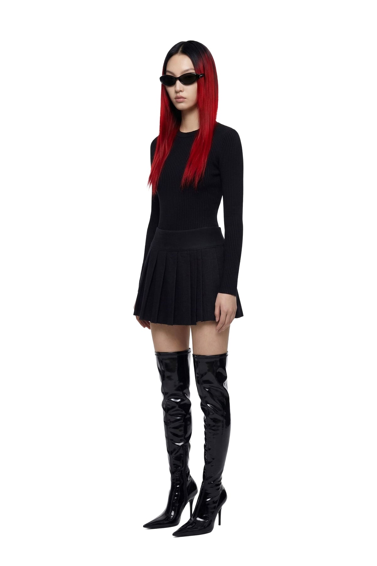 A model with long red hair wearing a black long-sleeve mini dress, black sunglasses, and shiny black over-the-knee boots, standing against a plain white background.