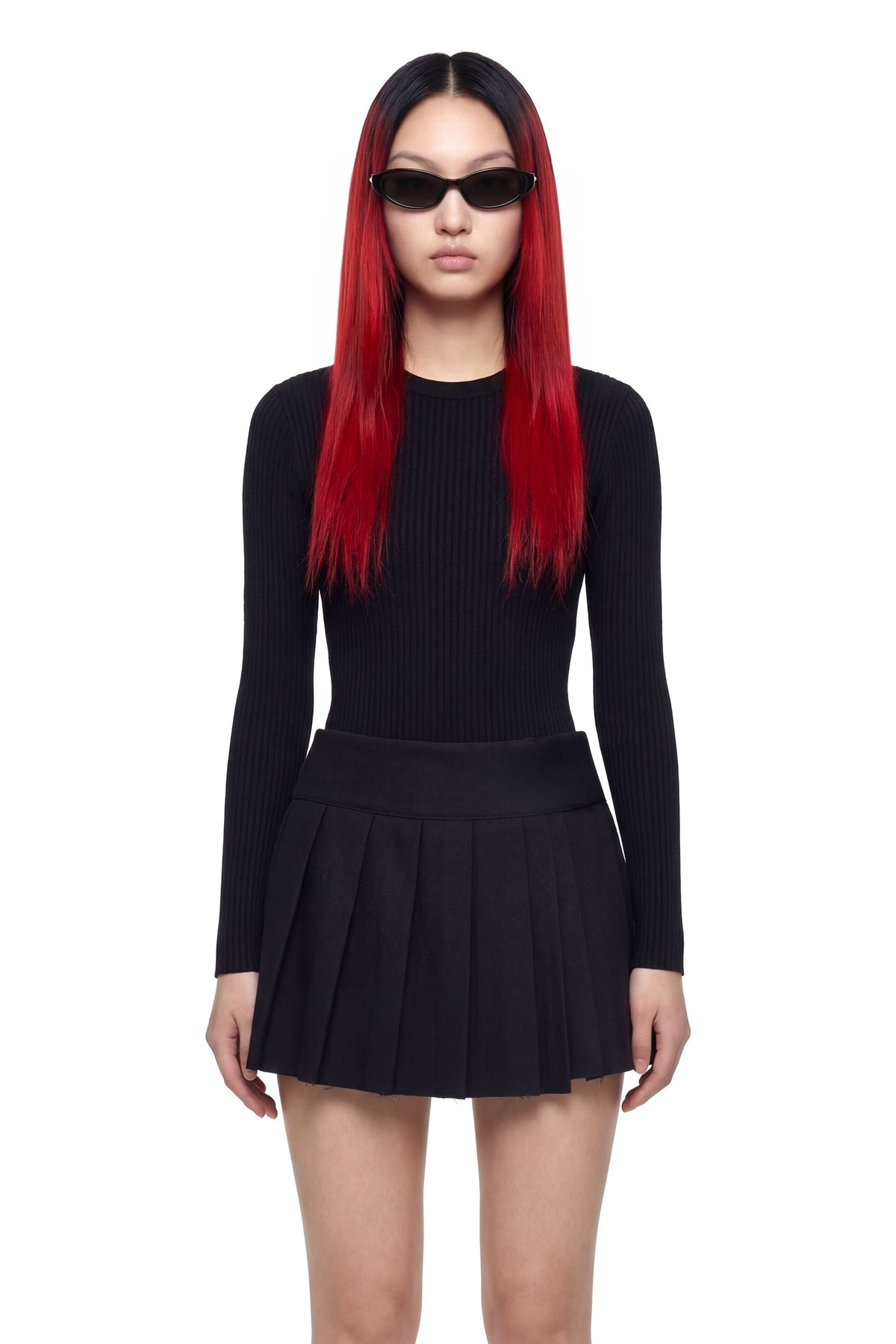 A model with long red hair and black sunglasses wearing a black ribbed long-sleeve top and a matching black pleated mini skirt, photographed against a plain white background.