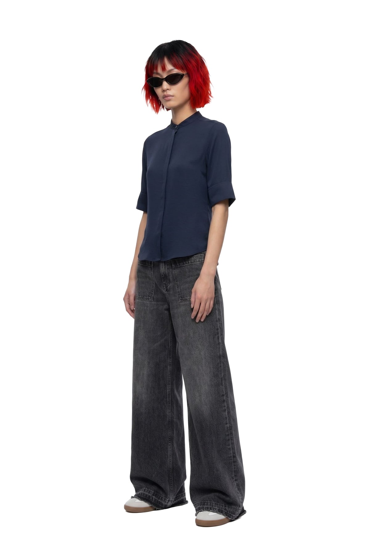 A model with short red hair and black sunglasses is shown from the side, wearing a dark navy blue short-sleeve button-up shirt, wide-leg faded gray jeans, and white sneakers against a plain white background.