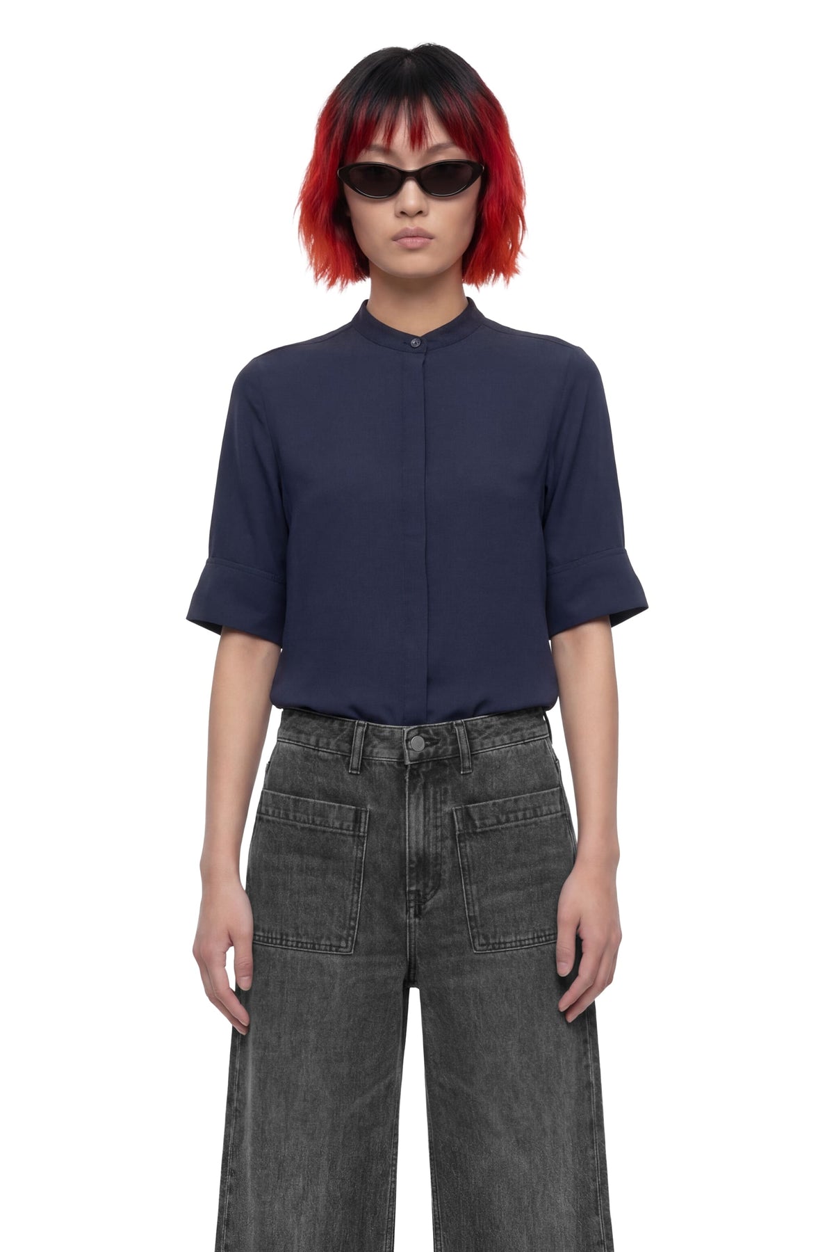 A model with a red and black bob haircut wearing a navy blue short-sleeve button-up shirt and dark gray wide-leg jeans, photographed against a solid white background.