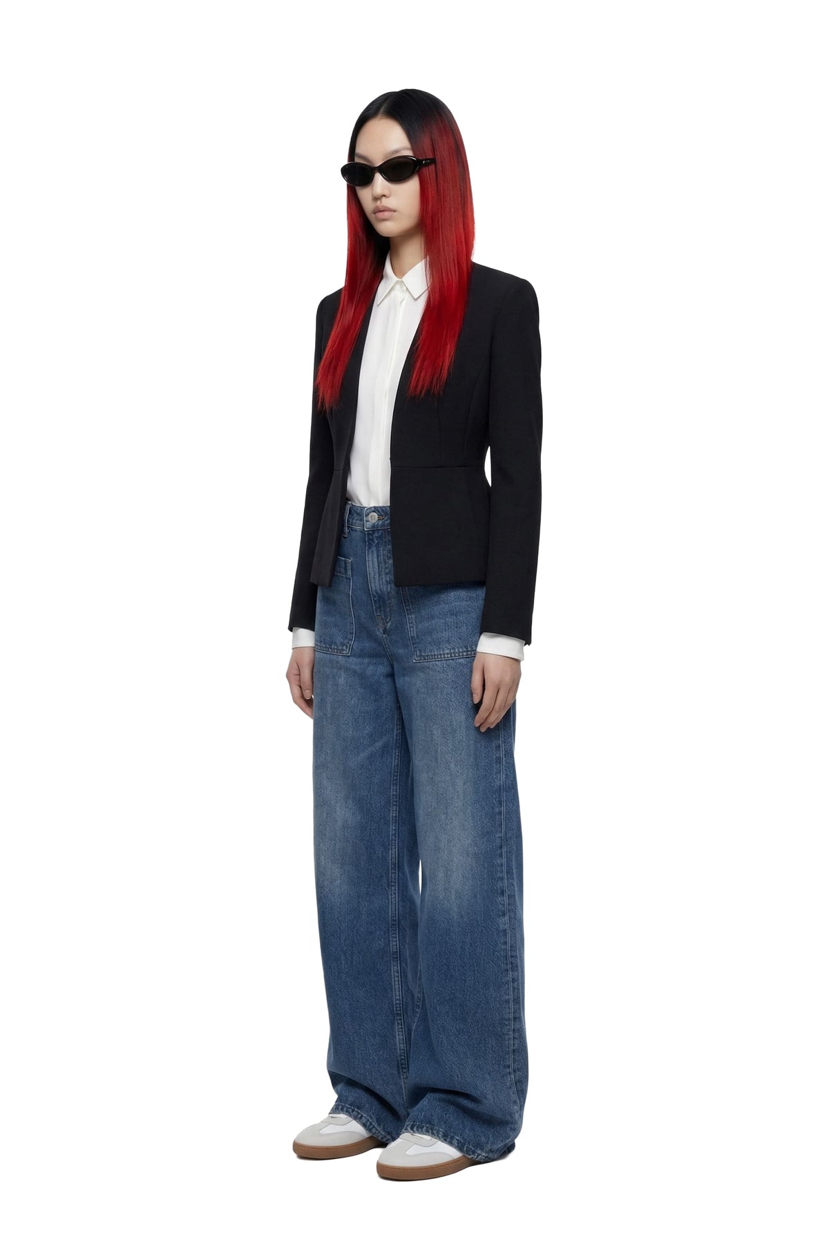 A model with long red hair wearing a black blazer, white collared shirt, wide-leg blue jeans, and black sunglasses, standing against a plain white background.