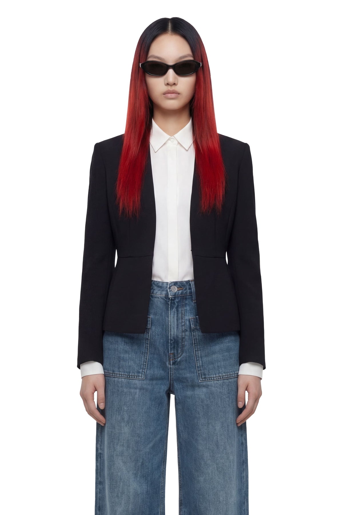 A model with long red hair wearing a black blazer, white collared shirt, and blue jeans against a plain white background.