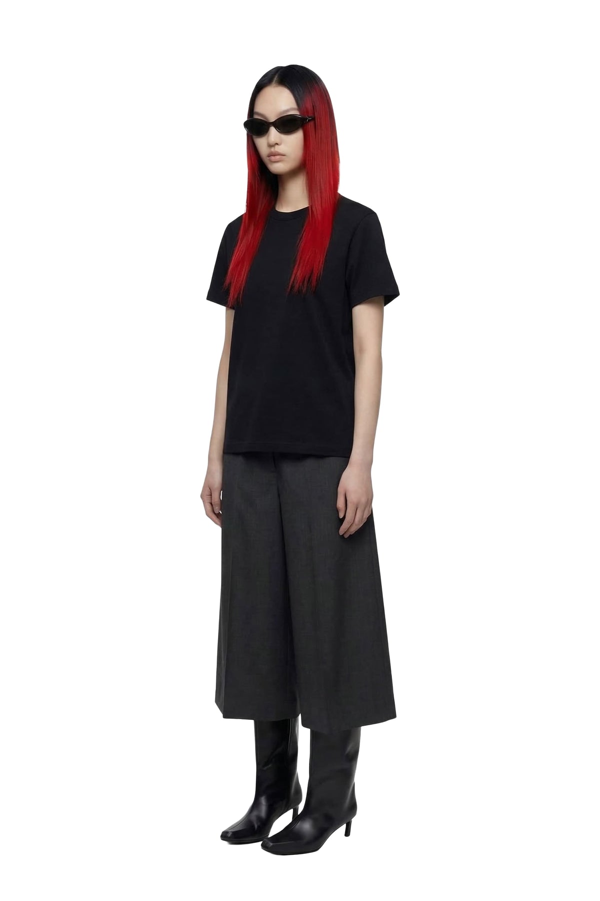 A model with long red hair and black sunglasses stands against a white background, wearing a black short-sleeve t-shirt, wide-leg black trousers, black knee-high boots, and black shoes.