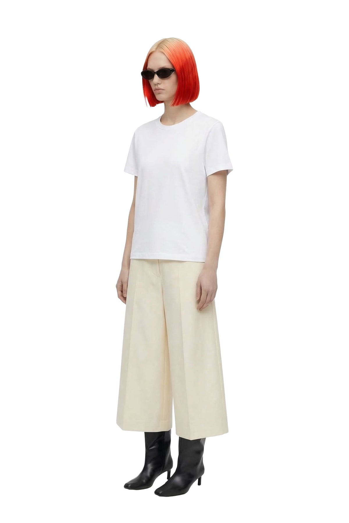A model with a vibrant orange-red bob haircut and sunglasses is shown from the side against a plain white background. She is wearing a white short-sleeve crewneck t-shirt, cream wide-leg cropped trousers, and black ankle boots.
