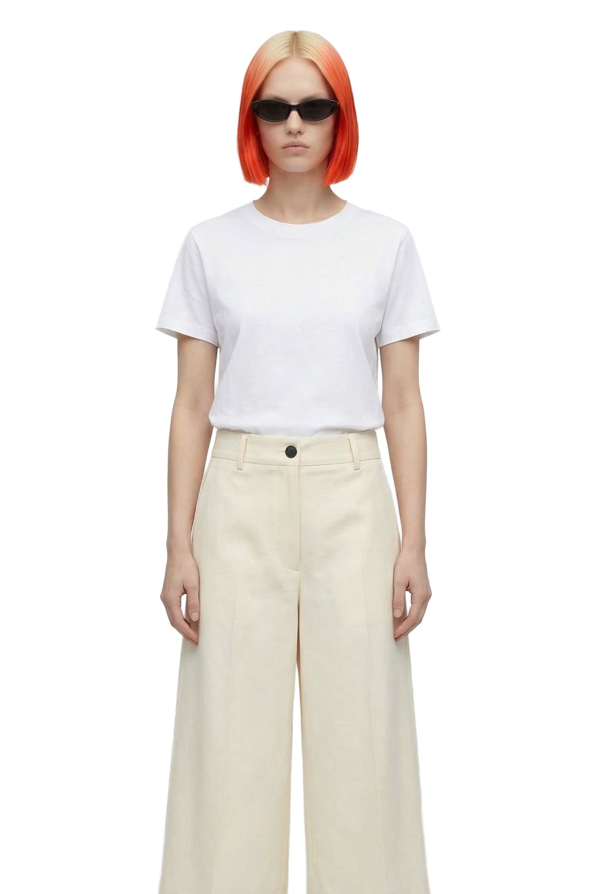 A model with bright orange hair and black sunglasses, wearing a white short-sleeve crewneck t-shirt and cream wide-leg trousers, photographed against a solid white background.
