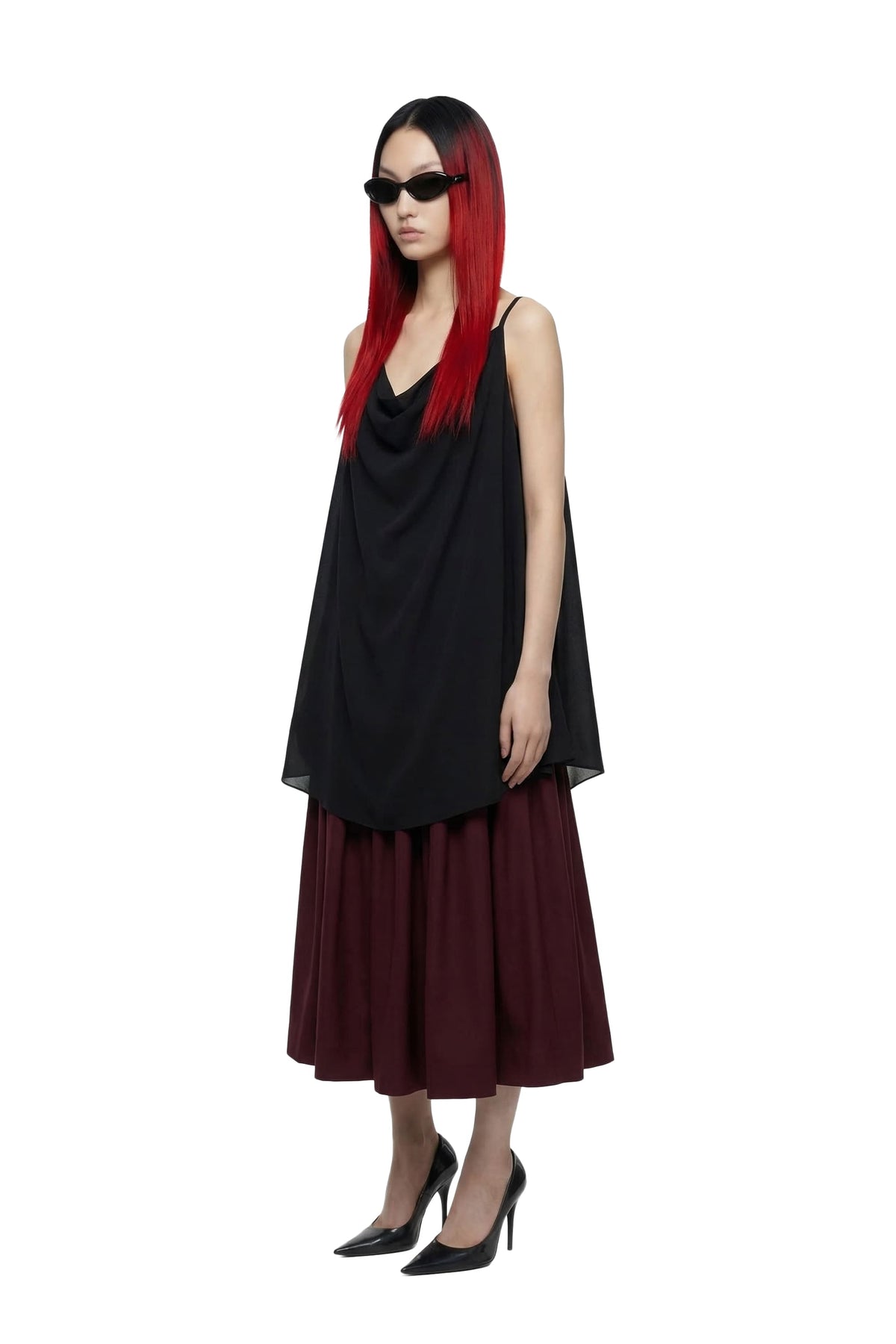 A model with long red hair and black sunglasses is wearing a black draped sleeveless top and a pleated dark red midi skirt, paired with black high heels, against a solid white background.