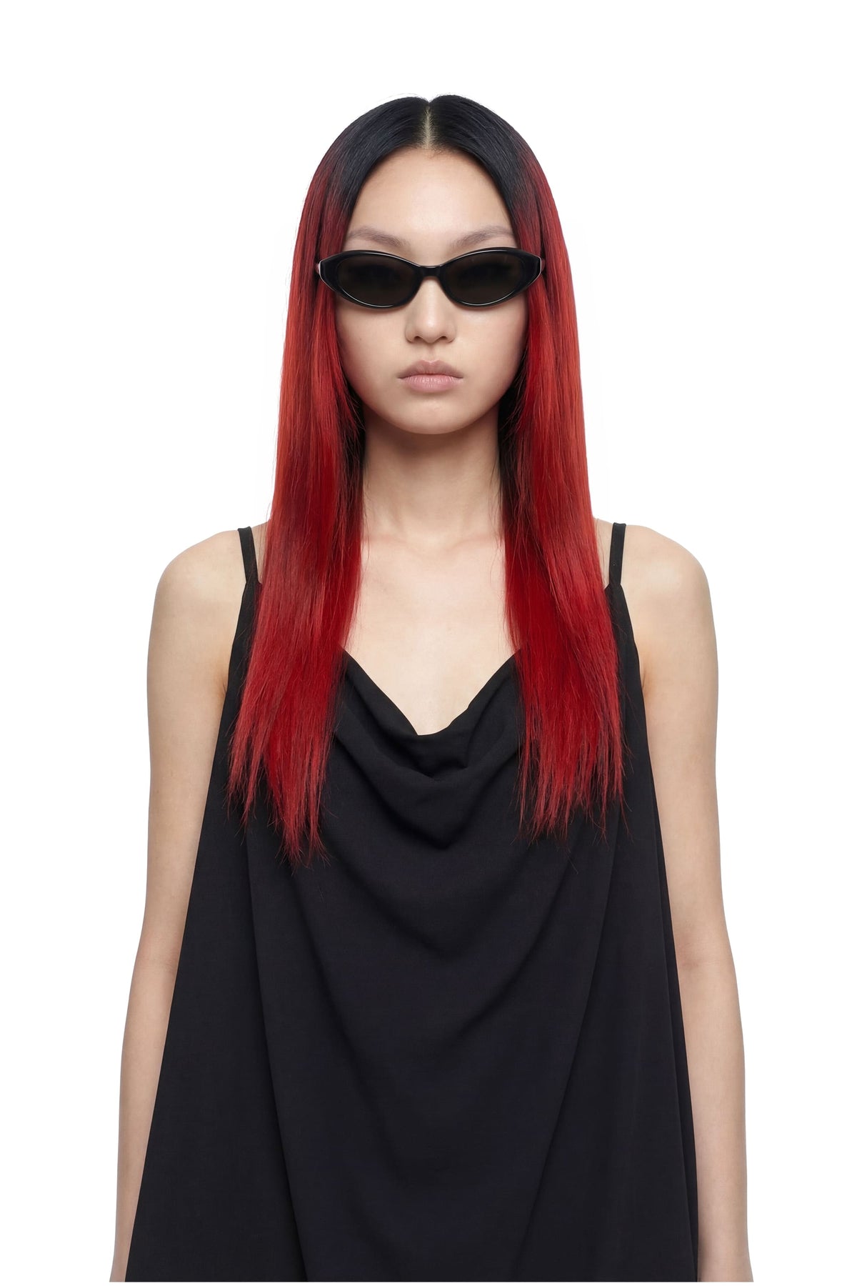 A model with long, straight black hair featuring vibrant red ombre ends, wearing a black draped sleeveless top and black cat-eye sunglasses, set against a plain white background.