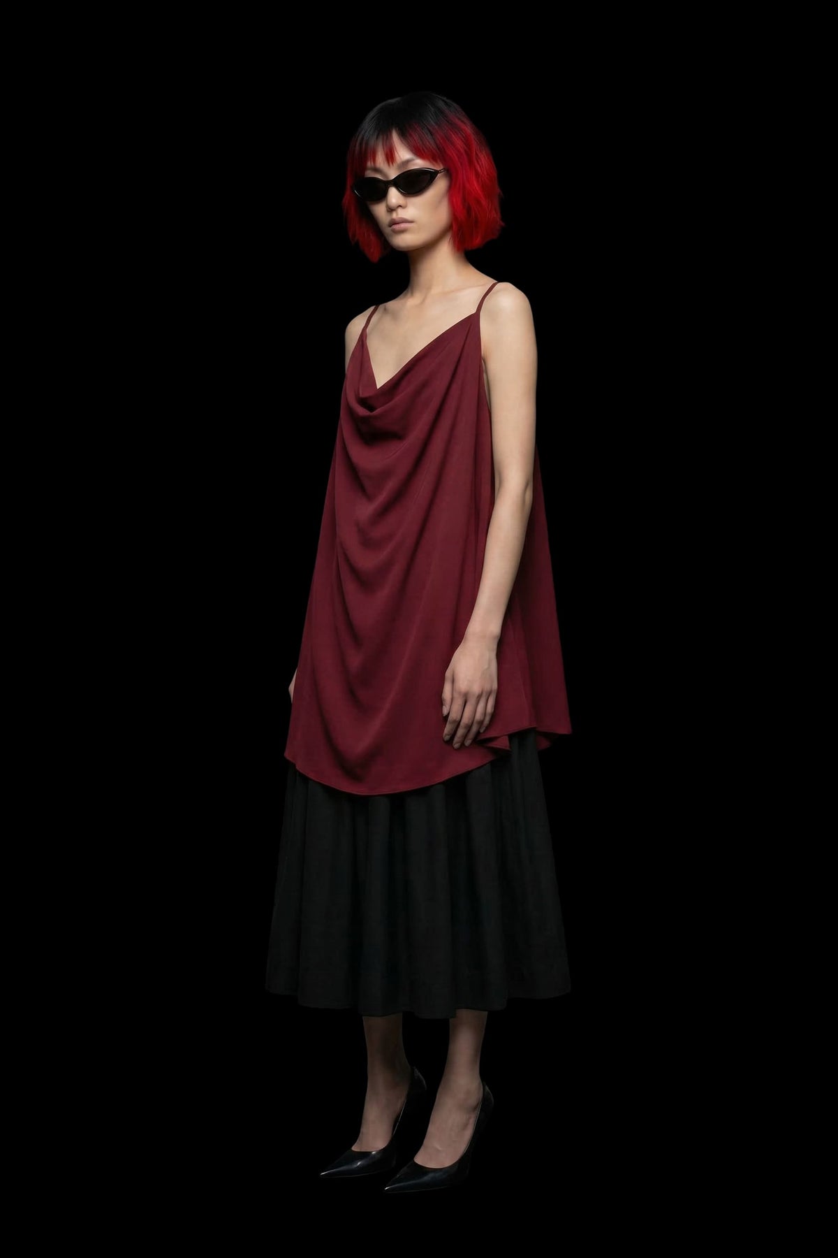 A model with short red hair and black sunglasses stands against a solid black background, wearing a dark red draped sleeveless top over a black midi skirt.