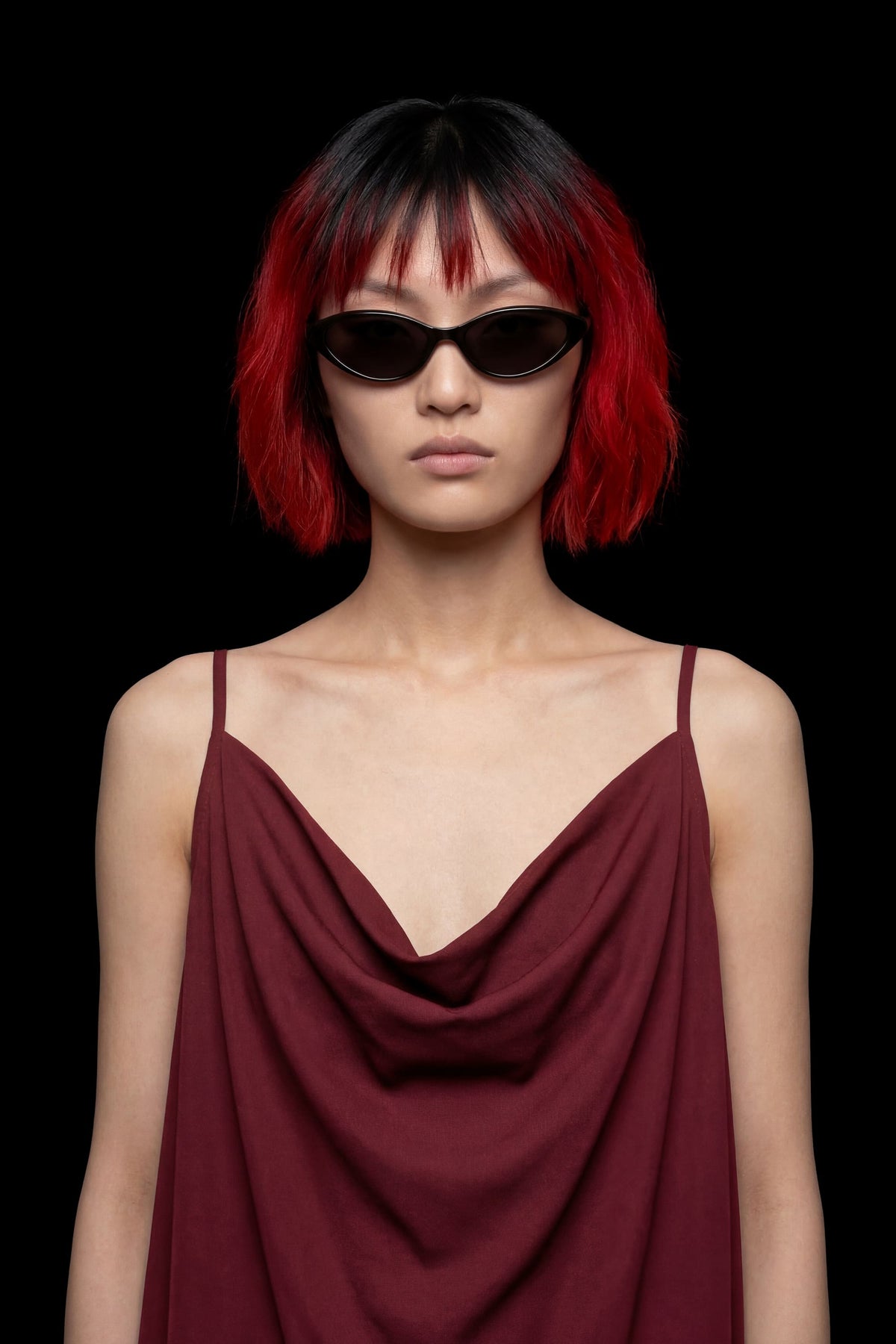 A model with a short red and black bob haircut wearing black sunglasses and a dark burgundy draped spaghetti-strap top, set against a solid black background.