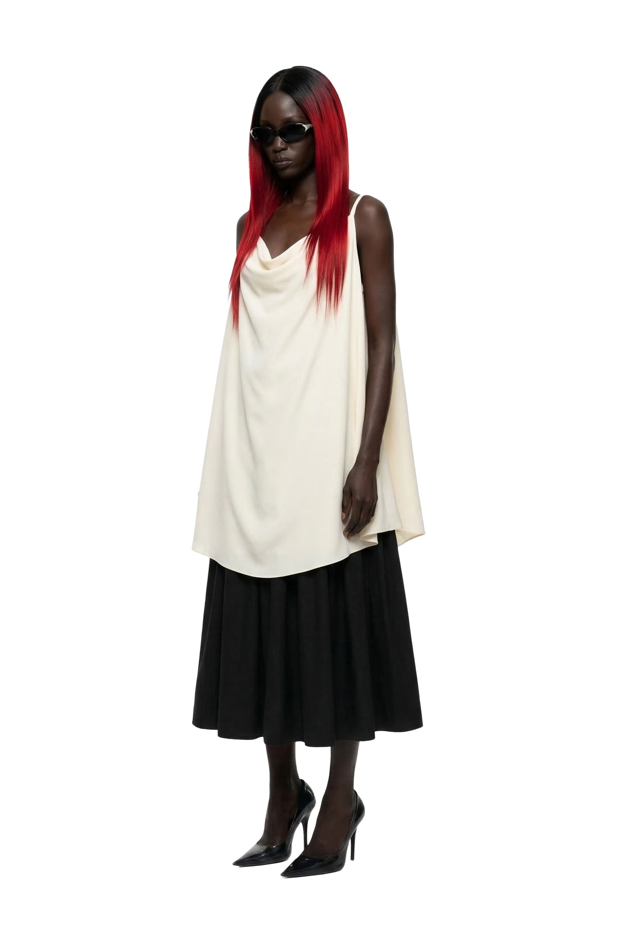 A model with long red hair wearing a sleeveless cream draped top, a black midi skirt, and black pointed-toe heels against a plain white background.