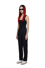 A model with long red hair and black sunglasses stands in profile wearing a black sleeveless top and black wide-leg trousers, paired with white sneakers, against a plain white background.