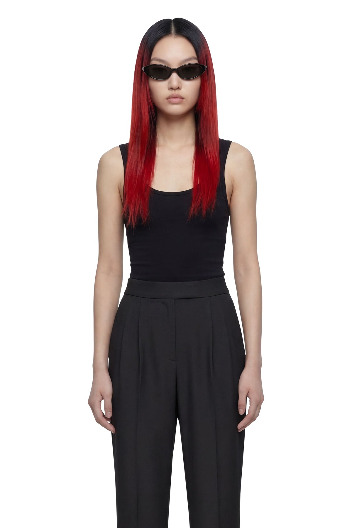 A model with long red hair and black sunglasses is wearing a black sleeveless top and black wide-leg trousers against a plain white background.