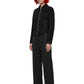 A model with short red hair and black sunglasses stands in a studio wearing a black zip-front jacket, black wide-leg trousers, and black shoes against a plain white background.