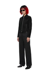 A model with short red hair and black sunglasses stands in a studio wearing a black zip-front jacket, black wide-leg trousers, and black shoes against a plain white background.