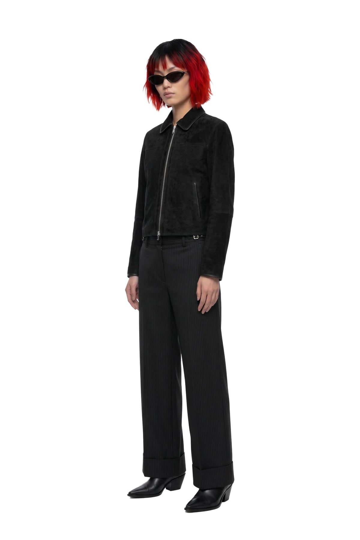 A model with short red hair and black sunglasses stands in a studio wearing a black zip-front jacket, black wide-leg trousers, and black shoes against a plain white background.
