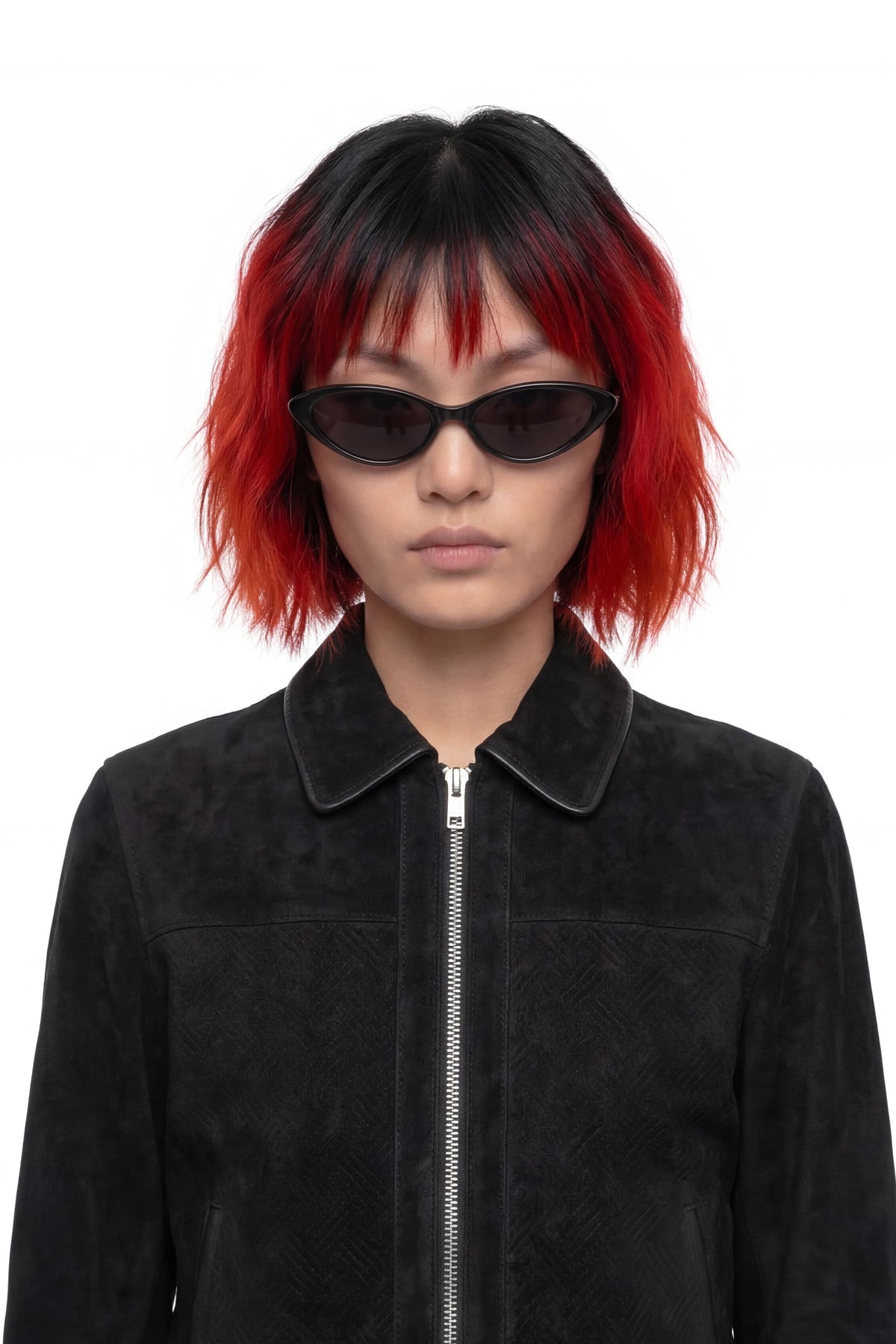 A model with a short black and vibrant red ombre wig wearing black cat-eye sunglasses and a black suede jacket, photographed against a solid white background.