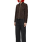 A model with vibrant orange hair and black sunglasses stands against a white background wearing a dark brown suede jacket, black wide-leg trousers, and black shoes.