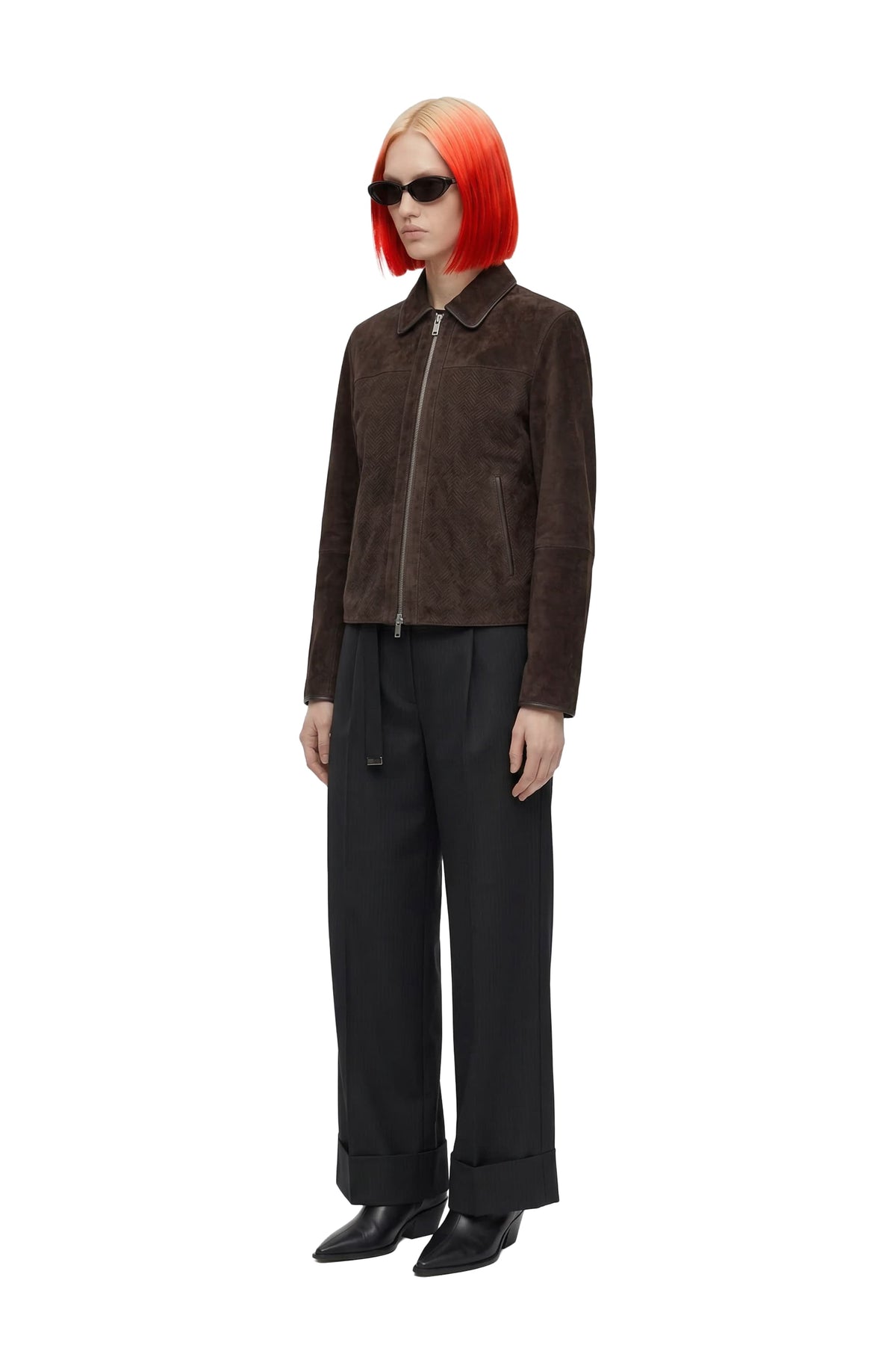 A model with vibrant orange hair and black sunglasses stands against a white background wearing a dark brown suede jacket, black wide-leg trousers, and black shoes.