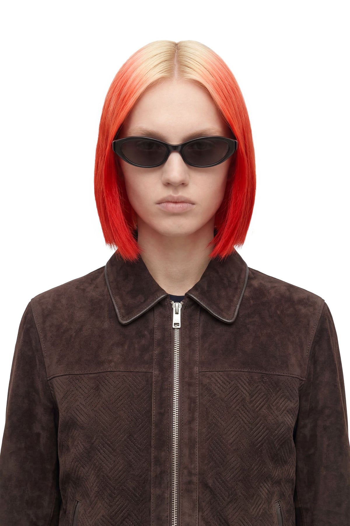 A model with a vibrant red-to-blonde ombre bob haircut wearing black sunglasses and a dark brown textured zip-up jacket, shot against a solid white background.