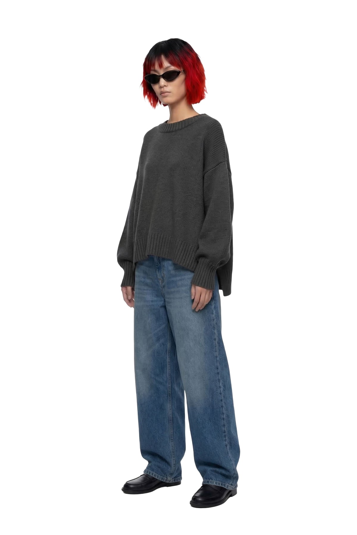 A model with a red and black bob haircut wearing a dark gray oversized knit sweater, wide-leg blue jeans, and black shoes, standing against a plain white background.