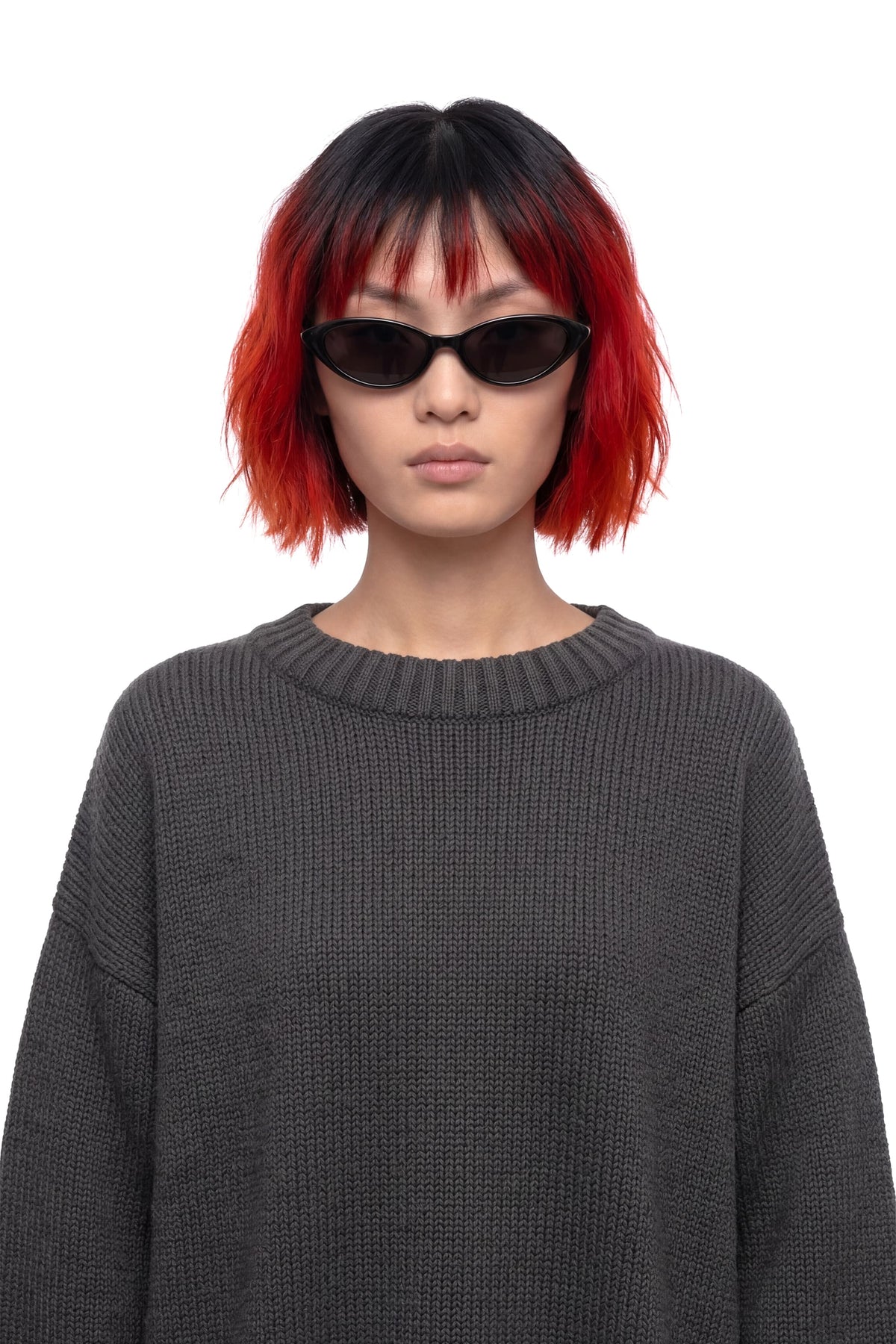 A model with a short black and red bob haircut wearing black cat-eye sunglasses and a dark gray ribbed knit sweater, photographed against a solid white background.