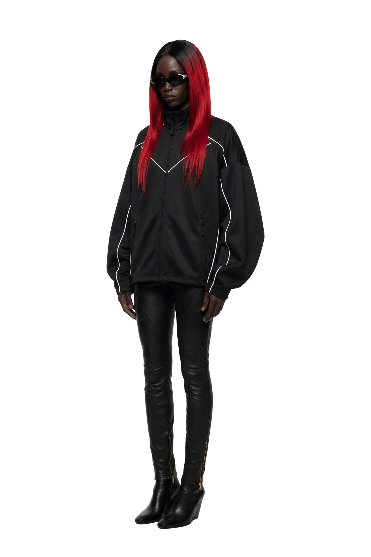 A model with long black and bright red hair wearing a black pullover sweatshirt with white piping details, black leather-look pants, and black ankle boots, set against a plain white background.