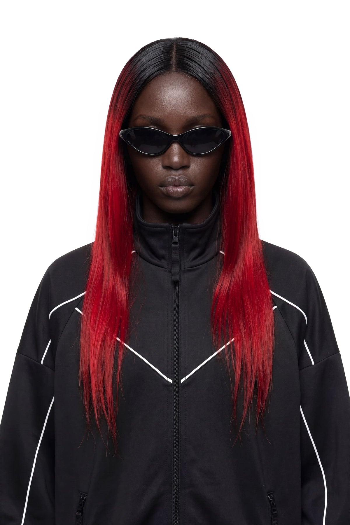 A model with long, straight black hair featuring vibrant red ombre ends, wearing black cat-eye sunglasses and a black zip-up jacket with white piping, set against a plain white background.