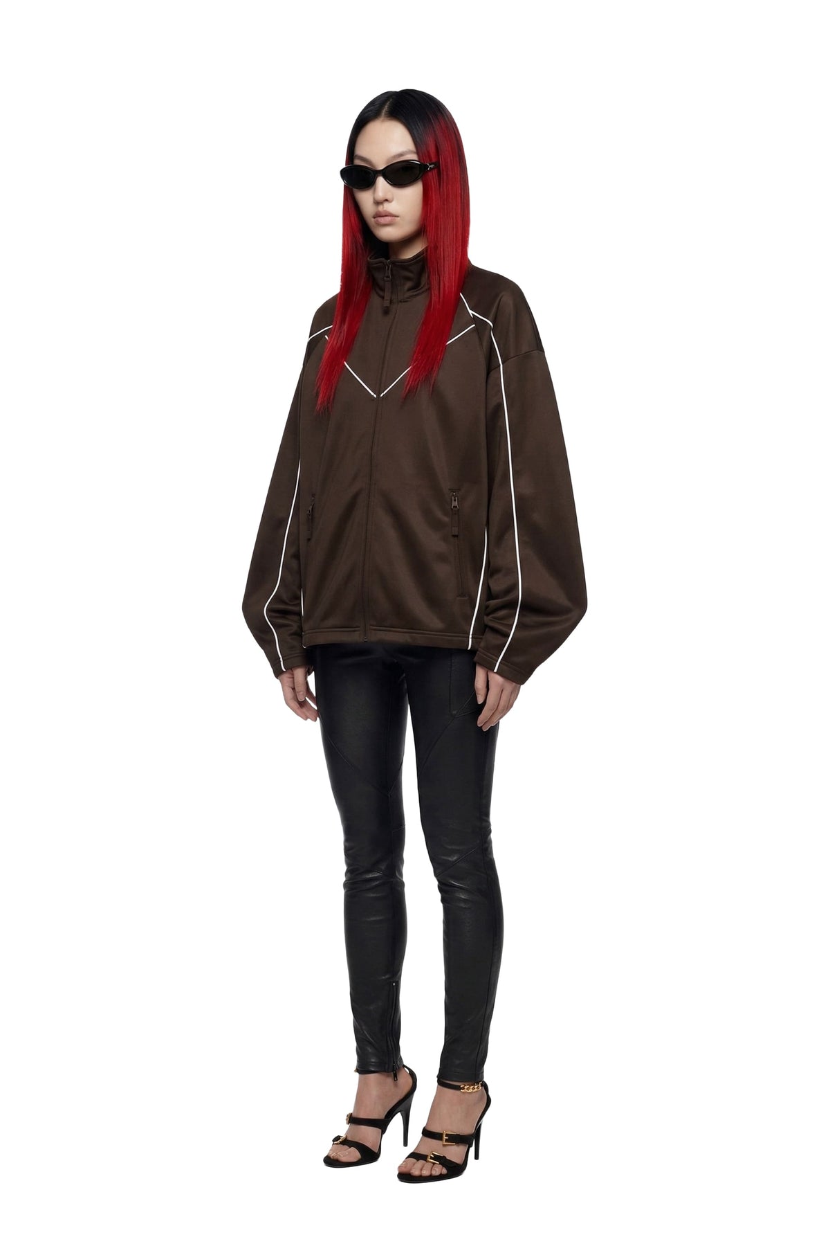 A model with long red hair and black sunglasses wearing a brown zip-up jacket with white piping, black skinny pants, and black high-heeled sandals, set against a plain white background.