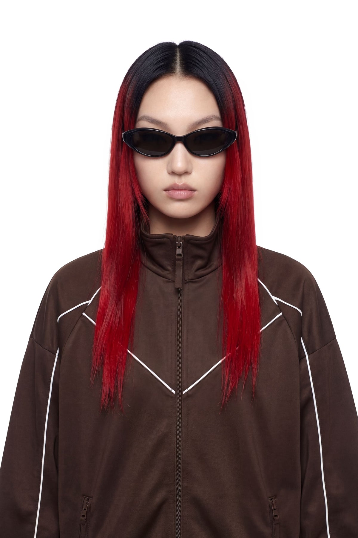 A model with long black hair featuring vibrant red ombre ends, wearing a dark brown zip-up jacket with white piping details and black cat-eye sunglasses, shot against a solid white background.