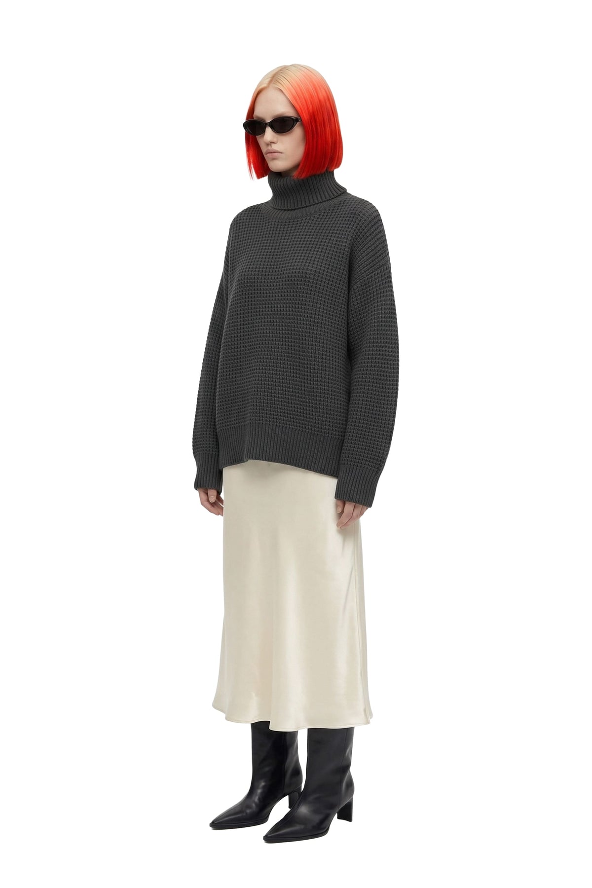 A model with a bright orange-red bob haircut and black sunglasses is shown wearing a dark gray textured knit turtleneck sweater, a cream satin midi skirt, and black pointed-toe boots against a plain white background.