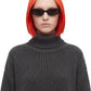 A model with a short, bright orange-red ombre bob haircut wearing black cat-eye sunglasses and a dark gray waffle-knit turtleneck sweater, set against a plain white background.