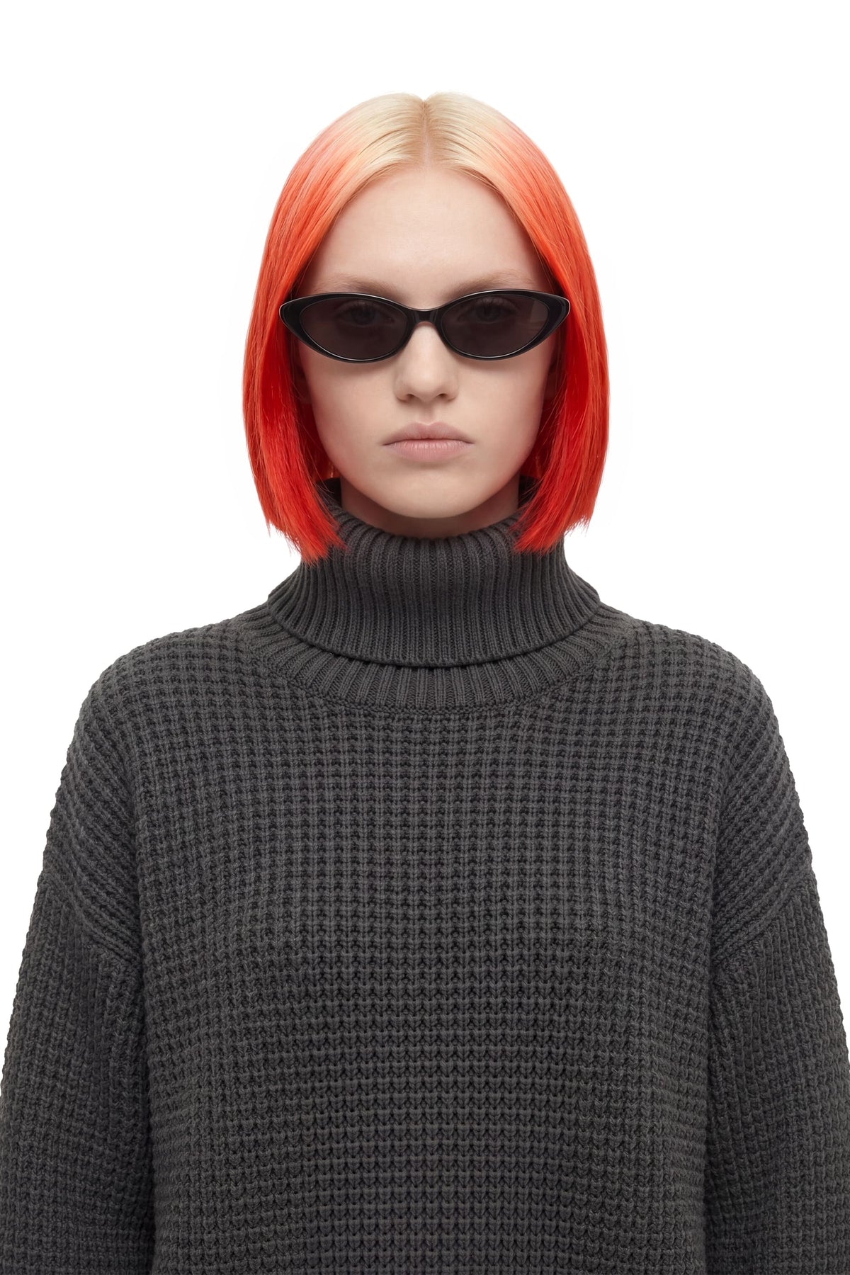 A model with a short, bright orange-red ombre bob haircut wearing black cat-eye sunglasses and a dark gray waffle-knit turtleneck sweater, set against a plain white background.