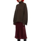 A model with a red and black bob haircut, wearing a dark brown oversized knit turtleneck sweater, a burgundy midi skirt, and black heeled boots, set against a plain white background.