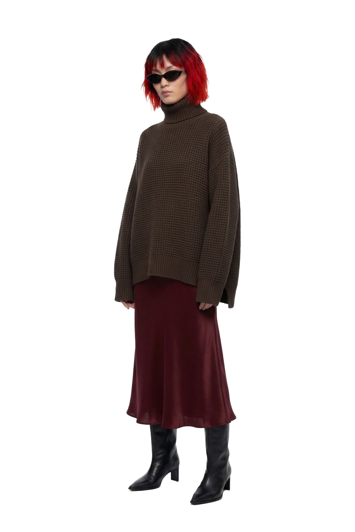 A model with a red and black bob haircut, wearing a dark brown oversized knit turtleneck sweater, a burgundy midi skirt, and black heeled boots, set against a plain white background.