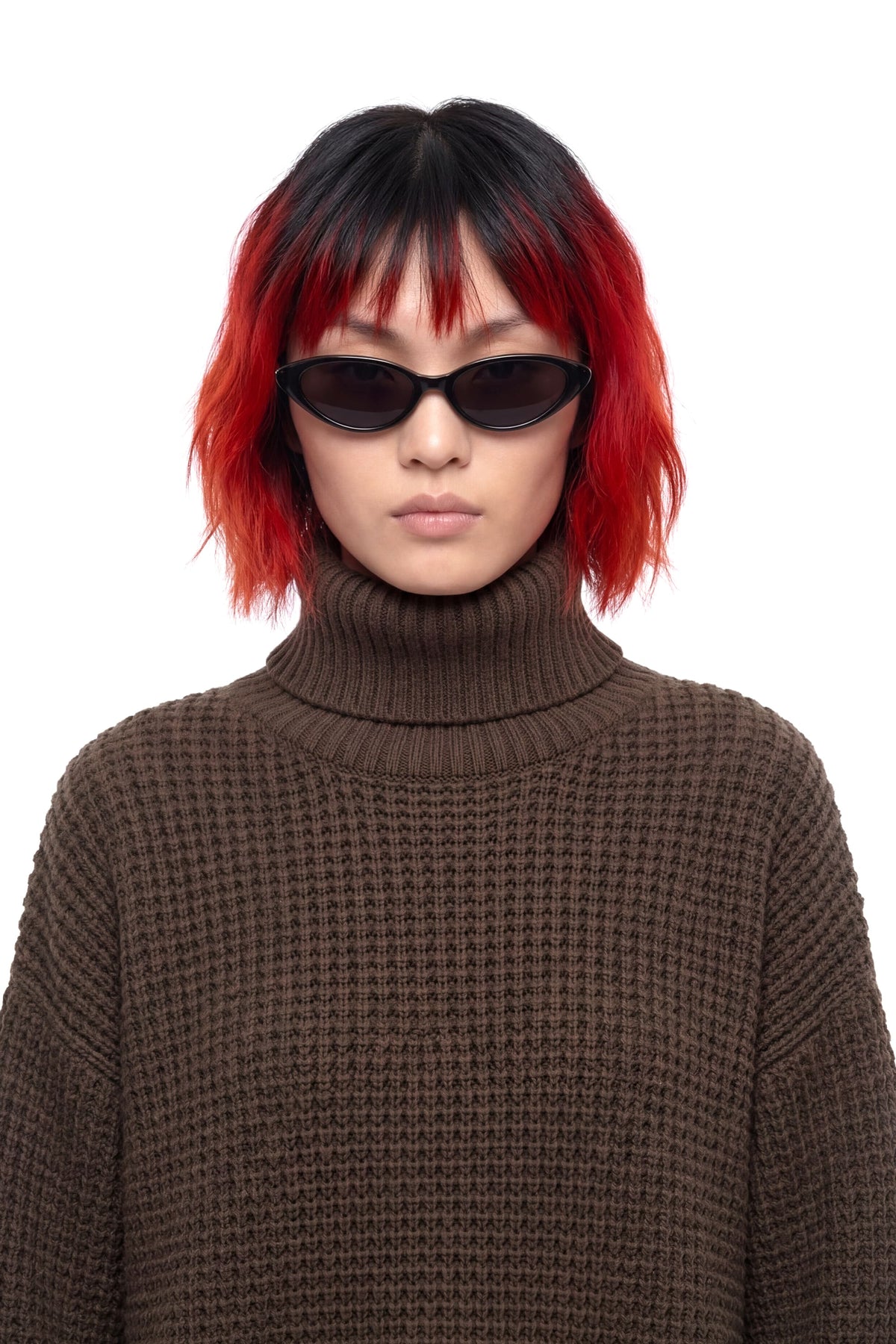 A model with a black and red ombre wig wearing a brown waffle-knit turtleneck sweater and black sunglasses, photographed against a solid white background.